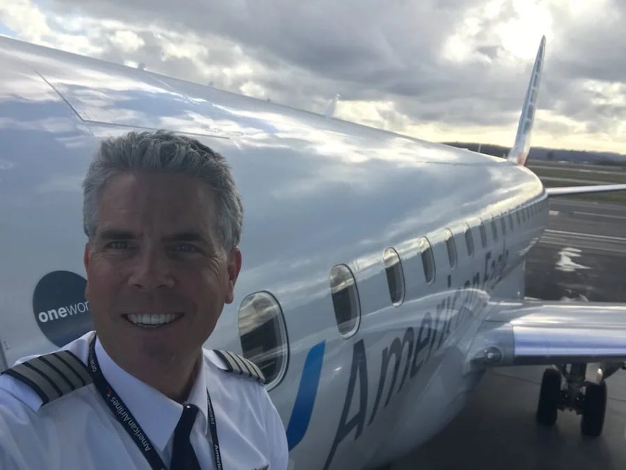 Robert Lawrence in airline captain's uniform, smiling beside an American Eagle jet on the ramp